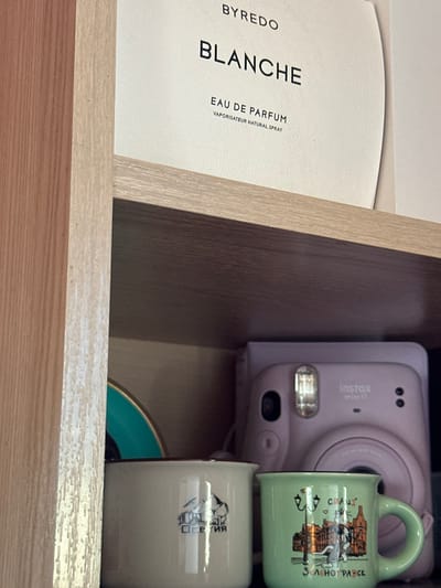 The image displays an indoor scene in the city of 3-Y Uchastok, Russia, featuring various objects arranged on and within wooden shelving. A light wood-grain shelf horizontally bisects the upper portion of the frame. Above this shelf, a white, curved-edge product package is visible, labeled with black text: "BYREDO", "BLANCHE", "EAU DE PARFUM", and "VAPORISATEUR NATURAL SPRAY". A vertical light wood-grain panel borders the left edge of the frame.
Below the shelf, positioned against a darker background, are three distinct items. On the far left, a partial view of a round, teal-colored object with a gold-colored rim is visible. Adjacent to it, a cream-colored mug with a dark brown rim displays a graphic of snow-capped mountains and the Cyrillic text "ОСЕТИЯ". To the right of the cream mug, a light green mug with a dark brown rim and handle features a multicolored illustration of an urban street scene, depicting buildings, a lamppost, and a white dog. This mug is inscribed with the Cyrillic word "ЗЕЛЕНОГРАВСК" at the bottom, and a partial word "CRAZ" accompanied by a heart symbol near the top. Behind these two mugs, a light purple Fujifilm Instax mini 11 instant camera is present, with its lens, reflective flash unit, and "instax mini 11" branding clearly visible. No individuals are present in the scene.
