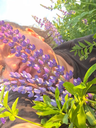 The image shows a close-up, low-angle shot of a person's face partially obscured by purple lupine flowers. The person's right eye is visible, along with their nose and mouth. They are wearing a dark-colored top. Lush green foliage and more lupine flowers are present in the background. The scene appears to be outdoors during daylight, with the sun casting shadows and highlighting the textures of the skin and petals. The location is identified as Doyna, Russia.