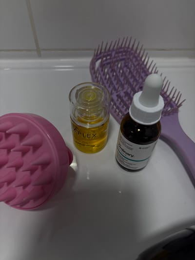 The image shows three items: a pink scalp massager with spiky bristles, a bottle of hair oil labeled "APPLEX" containing a clear, yellow liquid, and a bottle of dark liquid labeled "Ordinary". The items are on a white ceramic surface, which appears to be a bathroom counter. There is a purple hairbrush partially visible in the top right of the frame. The items are arranged in a triangle pattern. The image appears to have been taken in a bathroom. It is unknown who placed the items or what they are being used for. The location is a bathroom in Ramla, Israel, based on the provided coordinates.