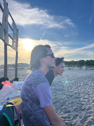 Two individuals are seated on a sandy beach in front of a low wooden structure. The person on the left has long, dark hair and is wearing a purple t-shirt with a patterned design. They are also wearing sunglasses. The person on the right has shorter, darker hair and is looking to their right. They are wearing a black t-shirt and sunglasses perched on their head. In the foreground, to the left of the individuals, are several bags, including a white tote bag with some items inside, a pink plastic bag, and a backpack with colorful straps. The background shows a beach area with more sand and a distant treeline. The sky is blue with scattered clouds and the sun is visible, casting a warm glow. The scene appears to be set in the city of Pistula, Montenegro.