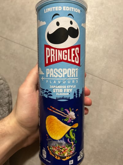 A hand is holding a can of limited edition Pringles Passport Flavours Japanese Style Stir Fry crisps. The can is blue and features the Pringles logo and an image of a wok of stir fry. The background appears to be a tiled floor. This does not occur in Nanterre, France.