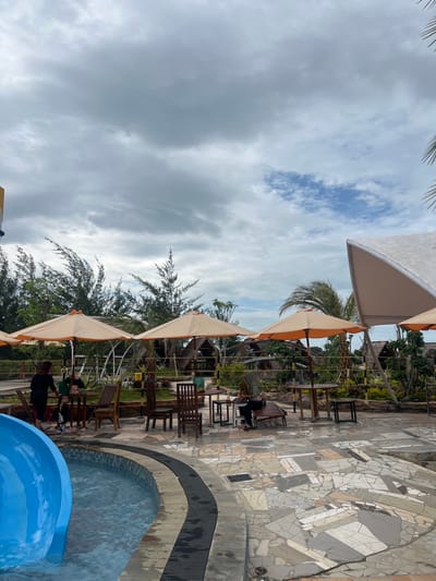 The image displays an outdoor recreational area in Juwana, Indonesia, under a predominantly overcast sky with a patch of blue visible in the upper right. In the lower left, a blue water slide empties into a swimming pool with a dark-tiled edge. The foreground and midground consist of a paved area featuring irregular, light-colored mosaic tiles. Four beige-orange open umbrellas are distributed across this area, providing shade for multiple wooden tables and chairs. On the left, an adult in a dark top stands near a table, interacting with a young child. Another child is partially visible nearby. On the right, an individual wearing a hijab is seated at a table under an umbrella. The background is dominated by dense green vegetation, including tall, slender trees and palm trees. Beyond the foliage, several thatched-roof huts or bungalows are visible, along with a large, light-colored structure featuring an angled roof on the far right.