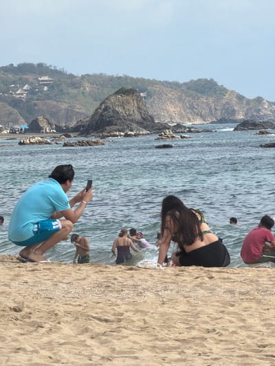 A sunny beach scene shows people enjoying the water. A man in a blue shirt and patterned shorts squats on the sand, holding up a phone as if taking a picture. Nearby, a woman with long, dark hair bends over towards the water. Further in, other individuals are wading, some partially submerged. A rocky cliff rises in the background, partially covered in vegetation. The sky is clear.