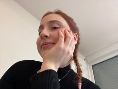 A female subject is depicted from a low-angle perspective, positioned against a plain, light-colored interior wall. Her gaze is directed upwards and slightly towards the viewer, with a subtle upward curve to her lips. She has reddish-brown hair styled in a braid, visible over her right shoulder, secured with a pink hair tie. Her right hand, with fingernails painted pink, is resting against her right cheek and chin. She is wearing a black ribbed turtleneck sweater and a thin silver chain necklace with a small, round pendant. To the far right of the frame, a portion of a white window frame and dark window glass are visible, indicating an indoor setting. The scene is illuminated by ambient, bright light. The image provides no identifiable features to determine a specific geographic location within Eschau, France, or elsewhere.