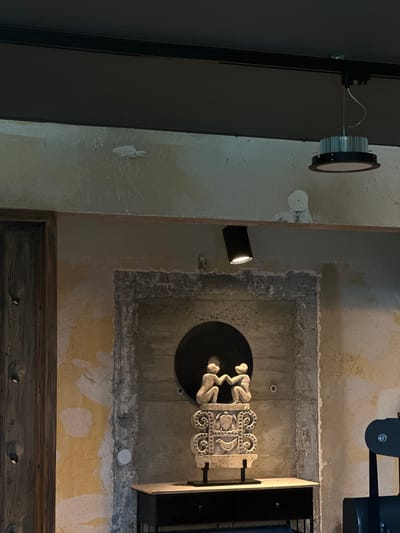 The image depicts an interior scene, likely in a contemporary or industrial-style space. The wall exhibits a distressed, unfinished texture with patches of yellowing paint or plaster. A dark wooden door is visible on the left. In the center, a rough, concrete-lined recess contains a circular black element. Resting on a dark console table in front of this recess is a light-colored carved stone sculpture. The sculpture features two figures at the top, appearing to shake hands or embrace, seated on an ornate pedestal with decorative carvings of birds and floral motifs. Above, a black spotlight is directed downwards, and a modern circular pendant light fixture hangs from a track on the ceiling. The overall impression is one of an art installation or a display in a gallery or modern home. The image is not recognizable as being in Bonn, Germany.