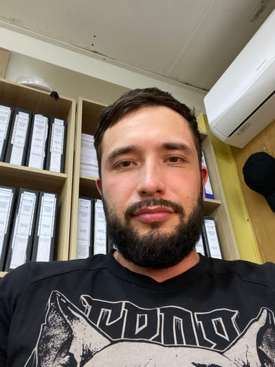 The image is a close-up selfie taken by a man with dark hair and a beard. He is wearing a black t-shirt with a graphic print. He is indoors, with shelves filled with binders behind him. An air conditioning unit is visible on the wall above the shelves. There is also a black hat hanging on the wall. The setting appears to be an office or storage area. The image does not contain any information indicating the location in the city of Luzhitsy, Russia.
