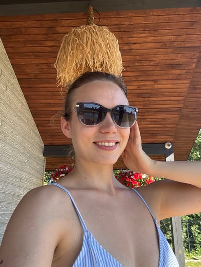 In this close-up, outdoor selfie, a young woman with light brown hair pulled back in a bun is wearing sunglasses and a blue and white striped bikini top. She is smiling and has her right hand resting on her cheek. The background shows a wooden ceiling, a hanging lamp made of straw-like material, and a wall with a textured finish. Red and white flowers are visible in the lower portion of the image, and in the reflection of her sunglasses, a body of water and trees can be seen. The lighting suggests it is daytime. The image does not provide any information that would place it in Rezekne, Latvia.