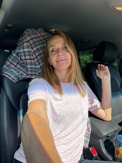 A woman with blonde hair and freckles is sitting in the driver's seat of a car. She is wearing a light pink t-shirt. She is looking at the camera and smiling. A plaid shirt is folded on the passenger seat. The car's interior is visible, including black leather seats and a center console. Sunlight streams in from the right side of the car, casting a shadow on the woman's arm. The image appears to have been taken during the daytime. The location is not Portovoe, Russia.