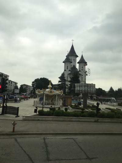 This detailed description pertains to an outdoor urban scene in Fălticeni, Romania, depicted under an overcast sky.
In the central background, a prominent religious edifice, likely a church or monastery, stands with a light-colored facade and multiple dark, conical spires, each topped with a cross. The tallest central spire displays what appears to be an electronic temperature reading of "+23°". Several evergreen trees are situated around the base of this structure.
The midground features a paved public square or park area. To the left, a decorative, enclosed carousel, primarily gold and white, is visible. Adjacent to it, a street runs parallel to a row of multi-story residential buildings. A traffic light showing a red signal is positioned on the left, next to a street sign partially legible as "REVOLUTIEI" and a pedestrian crossing indicator. Several vehicles, including red and black cars, are parked or driving on this street. Multiple individuals are present in the public square, some seated on benches, others walking. Their faces are indistinguishable due to distance.
In the center-right midground, a tall, modern sculptural monument rises, featuring a rectangular base section with sequential numbers "3" through "10" displayed on individual panels. Above this base, an intricate, multi-ringed spherical structure, resembling an atomic model, is mounted. Behind this monument, a small, light-blue fountain basin is partially visible. The public space is landscaped with trimmed green bushes and small trees, separated by concrete planters and paved walkways.
The immediate foreground is an asphalt road with visible cracks and patches, delineated by a concrete curb. A red fire hydrant is prominently located on the left side of this foreground road. No direct interactions or specific activities of the individuals are discernibly clear.