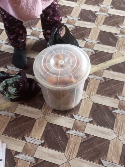 A plastic container with a clear lid sits on a floor with patterned tiles. The container appears to hold bread or other baked goods. Beside the container is a patterned scarf and a black plastic bag. A person stands in the frame, wearing black socks and patterned trousers. A pink garment covers their torso. A notebook or paper peeks into the frame's lower-left corner.