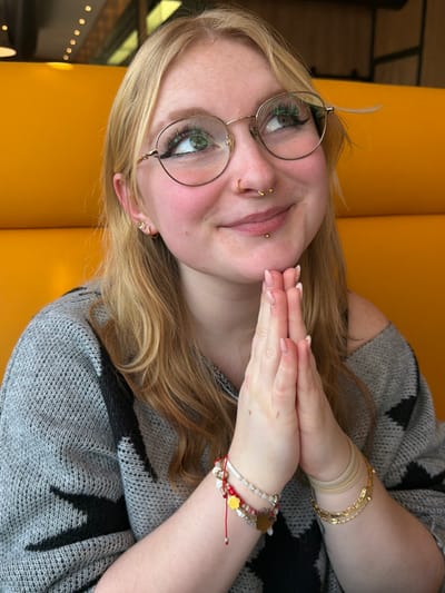 A blonde individual is depicted from the mid-torso up, seated against a bright yellow padded booth. The subject has shoulder-length blonde hair, gold-rimmed circular glasses, and green eye makeup. Facial piercings include a gold-colored hoop in the left nostril, a gold-colored horseshoe barbell in the septum, and a gold-colored barbell labret piercing. Small stud earrings are present in both earlobes. A subtle smile is evident, and the gaze is directed upwards and to the right. Both hands are clasped together, fingers interlocked, resting beneath the chin. The individual wears a grey knitted sweater with a black geometric pattern. The left wrist displays multiple bracelets, including beaded strands and a red string with a gold-colored charm. The right wrist features several thin, light-colored elastic bands and a gold-colored metallic bracelet. The fingernails are manicured with a light pink polish. The background shows the yellow booth seating and blurred overhead lighting fixtures. The location is Grevenmacher, Luxembourg.