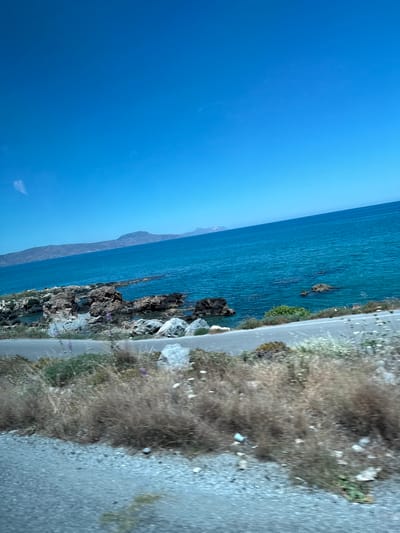 The image shows a coastal road in Rethymno, Greece, on a clear, sunny day. The sky is a vibrant, cloudless blue. The sea is a deep turquoise, with gentle waves breaking against a rocky shoreline. Mountains are visible in the distance, forming a hazy outline against the horizon. The road itself is paved and appears to be winding along the coast. On the side of the road, there is dry, scrubby vegetation, with some scattered rocks and wildflowers. The scene suggests a natural, undeveloped coastal area, likely a popular spot for scenic drives or exploring the natural beauty of the region.