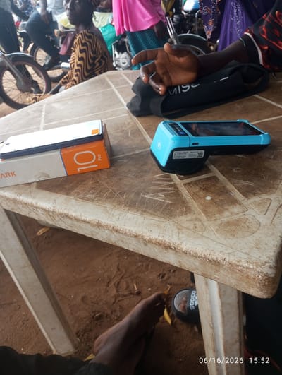 The image depicts a street scene in Dokan Tofa, Nigeria. A woman wearing a patterned dress sits on a wooden table, with a blue point-of-sale (POS) device and a smartphone on a box to her left. In the background, people are visible with motorcycles, suggesting a busy marketplace or street. The ground is made of red earth, and the table has a worn, plastic surface. The overall impression is one of everyday commerce and activity in a Nigerian setting.