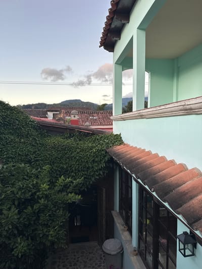 The image showcases the exteriors of buildings with varied architectural styles. One building has a pale blue facade with a white-trimmed roof. Adjacent to it is a building with terracotta roof tiles, heavily covered with climbing green foliage. A glimpse of a mountain range is visible in the background beneath a cloudy sky. The image is taken from a low angle, pointing upwards towards the structures.