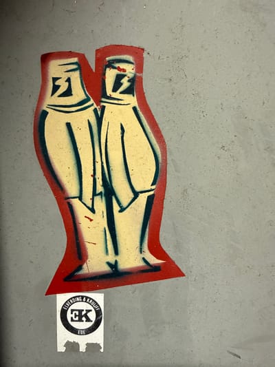 The image shows a stylized, red-outlined graphic with two horizontal shapes, possibly vases or figures, in a pale yellow color. They have dark blue markings. To the right is a small, circular sticker with black lettering on a white background, featuring "ELVERDING & KRUIFF" and "EK". All this is adhered to a gray surface.