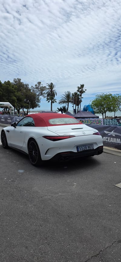 The image captures a white Mercedes-Benz AMG SL convertible with a red top parked on an asphalt road in Benicàssim, Spain. The car's license plate number is visible: 5528 NGS. Palm trees and other greenery line the background along with sky and clouds. Event banners are visible in the background for "OCEANMAN", suggesting an event is taking place in the area. The sky is overcast.