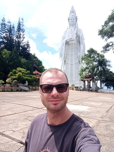 A man wearing sunglasses and a dark gray t-shirt takes a selfie in front of a large white statue of Guan Yin, the Goddess of Mercy, located in Nam Ban, Vietnam. The statue is tall and appears to be made of white stone or concrete. The man is positioned in the foreground, with the statue and its surrounding environment in the background.
The environment includes a paved courtyard with a cracked concrete surface. To the left of the statue, there are lush green trees and some smaller traditional Asian-style buildings with orange roofs. In the far distance, tall pine trees are visible against a bright blue sky with scattered clouds. The lighting suggests it is daytime, likely sunny. The statue's serene expression and posture are notable. The man's expression is neutral, and he is looking at the camera.