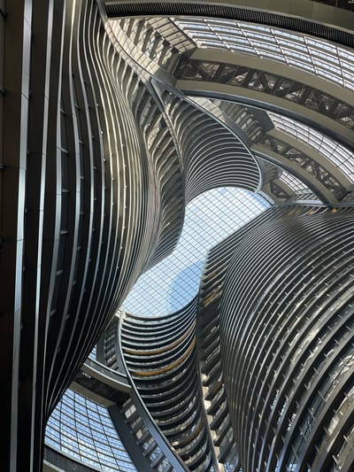 A low-angle shot within a modern architectural space, likely an atrium or lobby. The photograph is taken looking upwards. Curved, horizontally-layered structures rise and converge towards a grid-patterned glass ceiling that reveals the sky. The building materials appear to be metal and glass. The scene is well-lit, with light filtering through the ceiling. The location is Fengtai District, China. The building appears to be modern. No people are visible.