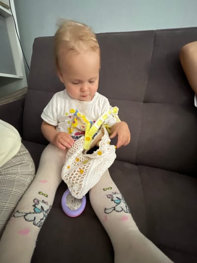 A young child with blonde hair, wearing a white t-shirt and patterned tights, sits on a dark brown couch holding a white knitted bag with yellow beads. The child is looking down into the bag, with one hand inside. A purple and pink toy is visible near the child's legs. The background shows a portion of a room with a white shelf on the left. The scene is indoors, suggesting a home environment.