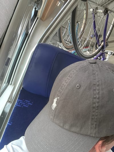 This media file captures an indoor scene, likely within a mode of public transportation such as a train, given the presence of blue upholstered seating and a window with a reflective metallic frame. A bicycle, partially visible with its purple frame and silver spokes, is mounted above and behind the seating, suggesting a bicycle-friendly transit system. In the foreground, a person's head is tilted down, wearing a dark gray baseball cap with a white embroidered logo of a horse. The person's ear is visible on the right side of the frame. The lighting appears to be natural, possibly indicating daytime, but the exact time is not discernible. The scene is dominated by muted colors, with the blue of the seats providing a contrast to the gray of the cap and the silver of the bicycle. No text is clearly visible within the frame. The overall impression is one of travel, with the bicycle indicating an active or recreational element to the journey. The orientation of the image, with the subjects appearing sideways, suggests it might have been taken while the person was seated and looking downwards or to the side.