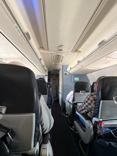 This media file captures the interior of an airplane cabin, rotated 90 degrees clockwise. The image looks down an aisle, with rows of passenger seats on both sides.
Several passengers are visible. In the middle-right foreground, a person is seated, wearing a brown and beige plaid shirt. A clear plastic water bottle is visible next to them. Further back, another passenger wears a light-colored (possibly pink) shirt. In the bottom-right foreground, the back of a black airplane seat is prominent, with a grey hooded sweatshirt draped over it, and another passenger partially visible. The seats are black with grey plastic components.
The overhead bins, typical of an aircraft, run along both sides of the aisle. They are light-colored, with handles and small information panels. Artificial cabin lighting illuminates the scene. The activity depicts passengers seated, likely during a flight or boarding/deplaning process.
Visible text on the overhead bins includes "23 CBAD" and "22 CBAO", indicating row numbers and seat letters. An emergency exit sign with the word "EXIT" and an arrow is partially visible further down the aisle. Small warning or instructional labels are also present on the cabin panels.
The overall setting is an aircraft cabin, consistent with travel, with the provided location context of Yagel, Israel, suggesting either the origin or destination of this journey. The time of day and weather are not discernible from within the cabin.