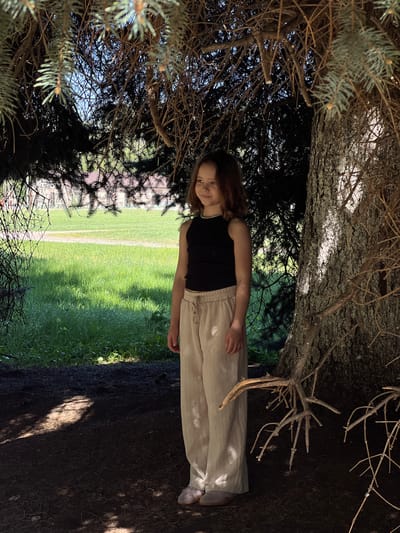 A girl stands beneath the canopy of a tree. The girl is wearing a black tank top and light tan pants. Part of a grassy lawn is visible in the background. The ground is dark and appears to be covered in dirt and tree debris. The location is likely Слобода, Russia.