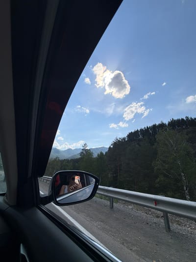 This is a view from inside a vehicle, looking out the passenger window towards a mountain landscape and a cloudy blue sky. The sun is partially obscured by a large cloud, creating a halo effect. In the foreground, a side-view mirror reflects a person in a white tank top, wearing sunglasses and holding a mobile phone. They appear to be taking a selfie. The road is gravelly, with a metal guardrail along the side. The mountains in the background are covered in dense green trees. The image does not provide any specific clues to identify the location as Turbaza Katun', Russia.