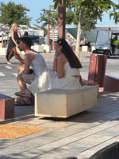 This is a medium-shot, eye-level photograph taken outdoors in a sunlit area that appears to be part of a promenade or public square in Eivissa, Spain, likely near a harbor, given the presence of a large ship in the background and what appears to be a golf cart used for transportation. In the foreground, a man in a white tank top and shorts is seated on a stone bench, holding an ice cream cone. Next to him, a woman in a white dress is also seated on the bench, facing away from the camera, with her hair styled with a flower clip. To the left, another woman in a black dress is walking away from the camera, also holding an ice cream cone. In the background, there is a large white cruise ship, a golf cart with a person wearing a high-visibility vest, and several trees. The ground is paved with stone slabs and there is a small square of orange gravel. The overall scene depicts a casual, outdoor setting with people enjoying themselves in warm weather.
