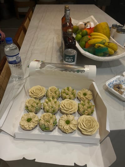 The image shows a white cardboard box containing 12 cupcakes, decorated with white frosting and green icing, shaped into different flower patterns, on a light-colored table. The table is set with a large white bowl containing various fruits, including limes, bananas, mangoes, and coconuts, and a can of "Selección Auto" next to it. The table is in a room with a beige tiled floor. A bottle of water and another bottle of alcohol are on the table, along with a white ceramic cup. This scene appears to be in a private residence or a catered event, located in Playa Herradura, Costa Rica. The specific location is a building with coordinates 9.658, -84.673.