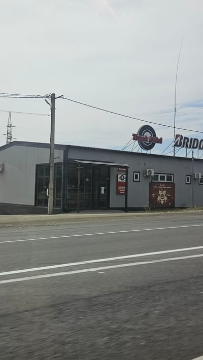 The image shows a building with a sign that reads "Truck Point" and "BRIDGESTONE". There is also a sign with a red star and some writing below it. The building appears to be a tire shop or service center. The scene is set in Verkhnebakanskiy, Russia. There are no people visible in the image. The environment is overcast with a road in the foreground.