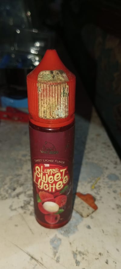 A bottle of Vapezoo "Lunar Sweet Lychee Flavor" e-liquid is shown. The bottle is red with a white and red label displaying the flavor name and lychee fruits. The cap is red, and a damaged, off-white sticker is partially attached to it. The sticker appears old and possibly moldy. The bottle is standing on a white, slightly dirty surface, next to an orange and white object.