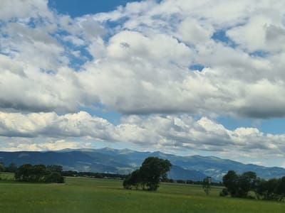 The image depicts a vast landscape with a prominent sky filled with dramatic clouds, interspersed with patches of blue. Below the sky, a range of blue-toned mountains recedes into the distance, layered with varying shades of green and blue. In the foreground, a lush green meadow stretches across the frame, dotted with scattered trees. To the left and center of the image, several large trees stand in the meadow, some with dense foliage. The scene appears to be a rural area in Ingering I, Austria, characterized by its expansive natural beauty. There are no people or animals visible in the image.