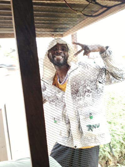 A medium shot captures a Black man wearing a white hooded jacket with a patterned design, a yellow undershirt, and black pants. He is smiling and giving a peace sign with his right hand. He appears to be in an outdoor, sheltered area, possibly a porch or covered patio, indicated by the corrugated metal roof and wooden beams visible. A net or mesh covers the foreground, obscuring parts of the man and the scene. Behind him, a blurred background suggests an outdoor environment with greenery and what might be a road with a vehicle. The overall lighting suggests daytime, with bright ambient light. The man's expression conveys a sense of happiness and casualness. The text "RNU" is visible on his jacket. The scene is set in Agwala, Nigeria.