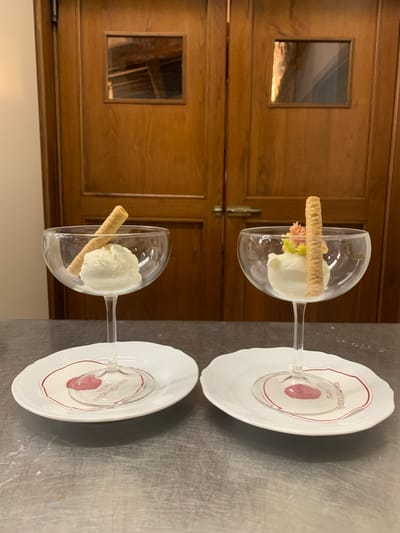 Two glasses of ice cream sit on white plates in front of a wooden door. The door has two square windows with reflective surfaces. Each glass contains a scoop of white ice cream, topped with a wafer stick. The left glass has only the ice cream and wafer. The right glass has a small garnish of pink and green fruit on top of the ice cream. The plates have a red border and a small drawing of a rose with text underneath. The scene appears to be indoors, likely in a restaurant or food establishment in Castiglione d'Orcia, Italy, given the text on the plate. The surface below the plates is a metallic, reflective countertop.