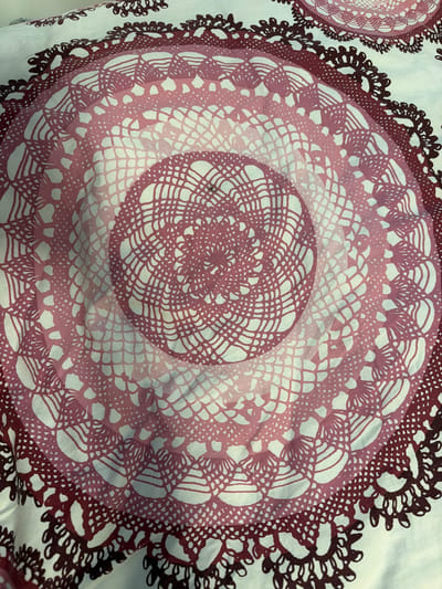 This is a close-up, overhead view of a fabric with a repeating circular, doily-like pattern. The pattern features concentric rings of intricate, lace-like designs. The colors transition from a deep reddish-brown at the outer edges to lighter shades of pink and white towards the center, creating a gradient effect. The design appears to be printed or dyed onto a white background. The fabric is slightly creased and appears to be a light material, possibly cotton or a blend. The lighting is neutral, suggesting an indoor setting without strong directional light. There are no people, animals, or specific setting details visible. The focus is entirely on the textile pattern itself.