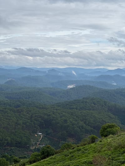 A high-angle, landscape photograph oriented vertically showcases a mountainous vista in Trường Thọ, Vietnam. On the right, a steep, grassy slope transitions into a dense forest with a mix of trees and foliage. A small clearing in the forest reveals a building complex with white roofs and structures. In the distance, the image features a series of hazy mountain ranges receding into the horizon. To the left of the mountains, the sky is filled with a thick layer of grey and white clouds.