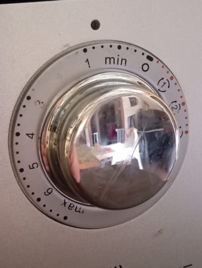 The image shows a close-up of a silver-colored timer dial on a kitchen appliance. The dial is circular with markings for minutes, ranging from "min" and "1" to "9" and "max." There are small black dots between the numbers, and red dots and circular arrow symbols at various points around the dial. The central part of the dial is a reflective silver knob. The background is a matte silver surface, likely part of the appliance. There are a few small dark specks on the surface of the appliance. The scene appears to be indoors. There is no indication of the city of Baden, Austria, in the image.