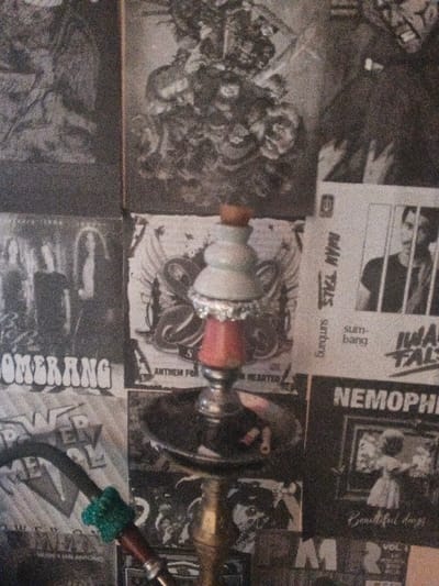This image depicts a close-up, cluttered interior scene in Dogsenang Gorphoog, Bhutan, dominated by a hookah positioned against a backdrop of numerous black and white posters and album covers.
In the foreground, a multi-component hookah stands centrally. Its upper bowl, covered with crinkled aluminum foil, is light-colored, possibly white or pale blue, and is topped with a small, reddish-brown cylindrical object. Below this, a reddish-orange colored column connects to a metallic, silver-toned base. A round, dark tray is situated below the red column, holding small, dark fragments, potentially ash or used shisha material. The hookah's stem descends into a brass or gold-colored section at the bottom, partially obscured. A black hose, accented with a vibrant green, textured fabric wrapping near its connection point, extends from the lower left side of the hookah.
The background consists of a wall entirely covered with monochrome (black and white) posters or album art. Visible elements include: a poster titled "BOOMERANG" with a band image on the lower left; "POWER METAL" below it; a partial title "AN ANTHEM FOR THE BROKEN HEARTED" directly behind the hookah base; "MAN TALES sunbang sumbang" on the upper right, featuring a man's face peering through bars; and "NEMOPHIL" with "Beautiful days" and an image of a child in a white dress on the lower right. Other posters display gothic, abstract, or heavy metal-themed imagery, including stylized skulls and band photographs. The overall impression is a personal space or establishment decorated with music-related paraphernalia, viewed under ambient lighting conditions, resulting in a somewhat grainy photographic texture.