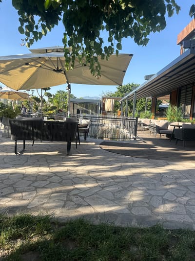 This image captures an outdoor seating area of a restaurant in Ashtarak, Armenia, under a clear blue sky. The foreground features a paved patio constructed with irregular-shaped, light-colored stones, interspersed with patches of green grass along the bottom edge. Sunlight casts dappled shadows across the patio.

Dominating the mid-ground are several large, light-colored umbrellas providing shade over a seating area. In the center, a dark-colored, woven bench with cushions faces away from the viewer. Behind the bench, a black metal railing encloses a body of water, likely a pond or small canal, reflecting the sky and surrounding greenery.

Beyond the water, the restaurant's outdoor seating continues. A wooden deck area hosts several upholstered sofas and chairs, suggesting a dining or lounge space. The restaurant building itself, visible in the background to the right, has a modern design with wooden accents and large windows. Green trees and manicured bushes are scattered throughout the scene, contributing to a pleasant, landscaped environment. In the far distance, mountains are faintly visible against the horizon. No people are prominently featured in the image, though a distant figure can be seen near the umbrellas.
