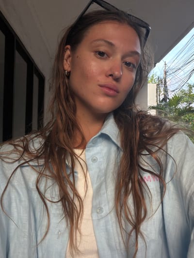 A young woman with long, wavy, reddish-brown hair poses for a selfie. She wears a light blue button-down shirt over a white top. A pair of sunglasses rest on top of her head. Her skin tone appears light with some visible freckles. She has pierced ears. The background shows a wall, overhead power lines, a glimpse of greenery, and a building. Given the prompt, the location can be inferred to be Kerobokan Kelod, Indonesia.