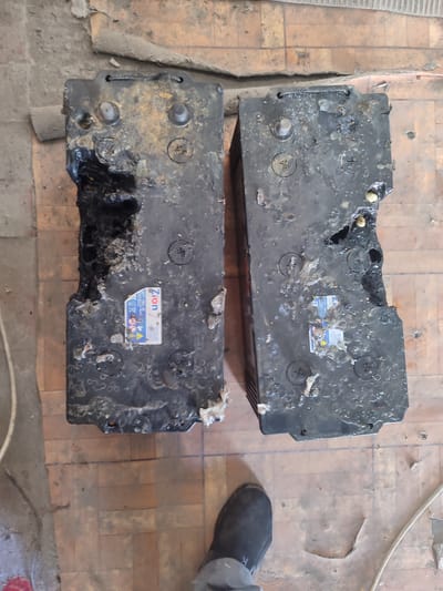 Two heavily damaged car batteries are lying on a floor made of orange and brown tiles. The batteries are black and appear to be covered in a sticky, white residue. One battery is missing a significant portion of its side, revealing internal components. The other battery also shows signs of damage and melting. Both batteries have labels on them, but the text is difficult to read due to the damage and residue. One label, partially visible, has the brand name "Zion" and some text below it. The floor is dirty and appears to be in disrepair, with some tiles cracked or missing. In the lower right corner of the image, a person's foot wearing a dark shoe is visible, suggesting a person was present when the photograph was taken. The scene appears to be indoors, possibly a garage or workshop, given the presence of construction materials and debris in the background. The lighting suggests it could be daytime. The overall impression is one of damage and neglect.