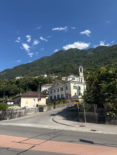 The image captures a street view in Magadino, Switzerland, with a mountain slope covered in dense green trees dominating the background. The sky is a clear blue with scattered white clouds. In the foreground, a paved road curves through the scene. To the left of the road, a low stone wall borders a property adorned with white flowers.
Further up the road, a pale yellow building with green shutters stands prominently. This building features a clock tower with a bell, suggesting it might be a church or a municipal building. Next to it, another building with a similar architectural style is visible. Several road signs, including traffic restrictions and directional signs, are posted along the roadside. Palm trees and other lush vegetation are scattered throughout the scene, contributing to the Mediterranean feel. Several other buildings, some residential and some appearing to be commercial, are nestled on the hillside behind the main structures. The overall impression is a sunny day in a picturesque Swiss town.