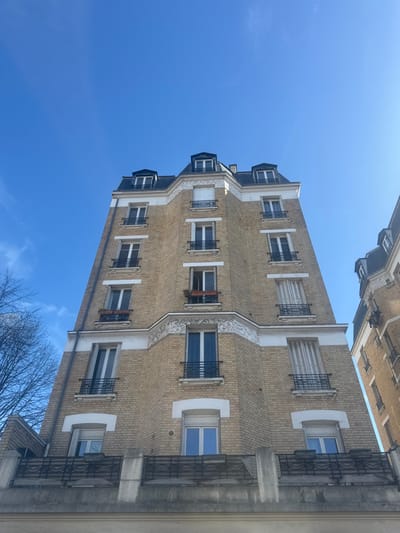 A multi-story building in Asnières-sur-Seine, France, is depicted from a low angle against a clear blue sky with minimal wispy clouds. The facade is primarily composed of light beige brickwork, accented with white horizontal bands and ornate white decorative elements, particularly on a central projecting bay. Numerous windows with dark frames are visible; many feature black wrought-iron balconies or balustrades. A prominent concrete balcony with dark, wavy-patterned metal railings spans the width of the lower visible section. Above this, on the third floor from the bottom (left side), a window is partially open and contains a brown planter box. A fourth-floor window on the central bay also displays a red-brown planter box. The building is topped with a dark mansard roof featuring multiple dormer windows. A portion of a similar adjacent building is visible on the far right. Bare deciduous tree branches are visible on the far left. No people are present, and no actions or interactions are observed.
