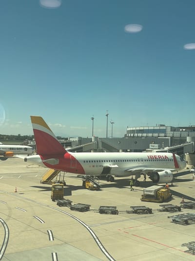 This media file captures an outdoor scene at an airport, likely Schwechat, Austria, on a clear, sunny day. The primary subject is an Iberia airplane parked at a gate, connected to the terminal building via a jet bridge. The aircraft is predominantly white with red and yellow accents on its tail and fuselage, and the "IBERIA" logo is clearly visible in large red lettering. The registration "EC-NTA" is also visible on the plane.
Several objects associated with airport operations are present on the tarmac. These include baggage carts, some filled with luggage, and ground support equipment like a yellow baggage loader. A few individuals, presumably airport personnel, are visible near the aircraft and equipment. One person in a yellow vest is near the baggage loader, and another in a high-visibility vest is standing on the tarmac.
The scene appears to be set during daylight hours, with strong shadows cast by the aircraft and the terminal building, indicating it's likely midday or early afternoon. The sky is a vibrant blue, suggesting fair weather. The terminal building, with its modern architecture and large windows, serves as a backdrop to the parked aircraft. The overall impression is one of a typical airport operation, with an aircraft being serviced or prepared for its next flight.