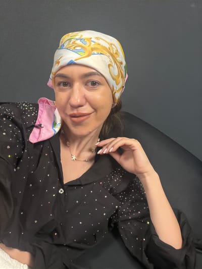 A woman is pictured in a close-up shot, posed against a dark, possibly upholstered surface. She is wearing a black, long-sleeved garment adorned with numerous small, sparkling embellishments, giving it a glamorous appearance. A colorful silk scarf with a swirling gold and blue pattern is tied around her head, with a pink edge visible near her collarbone. She is also wearing a delicate necklace featuring small charms. Her expression is a gentle smile, and her eyes are looking towards the camera. Her right hand is gracefully positioned near her face, with manicured fingernails. The lighting suggests an indoor setting, likely during the daytime, with soft illumination highlighting her features and the reflective details on her clothing. There is no visible text in the image.