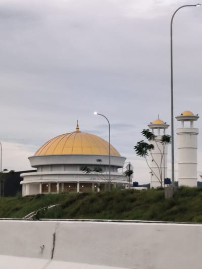 The image captures a modern mosque or Islamic religious complex, viewed from a roadside perspective under an overcast sky. In the mid-ground, a central building features a large, striking golden dome atop a circular white structure with an open, perhaps colonnaded, base. To the right, two slender white towers, resembling minarets, rise, each topped with a smaller golden dome.
The buildings are nestled on a vibrant green, grassy slope. Sparse trees with light green leaves are scattered around the structures and along the slope. In the foreground, a white concrete road barrier occupies the lower portion of the frame, indicating the vantage point is from a road or highway.
Several streetlights are visible. A tall, dark grey streetlight with an illuminated lamp stands prominently on the far right. Another streetlight with a glowing lamp is positioned closer to the main golden dome on the left.
The sky is entirely overcast, casting a soft, diffused light over the scene. There are no people visible, and no discernible activity is taking place. The overall impression is one of contemporary religious architecture set against a natural, green landscape. The visible streetlights being on could suggest either early morning, late afternoon, or simply very cloudy conditions. No text is visible in the image. This architectural style and setting are consistent with the provided location context of Cotkareung, Indonesia.