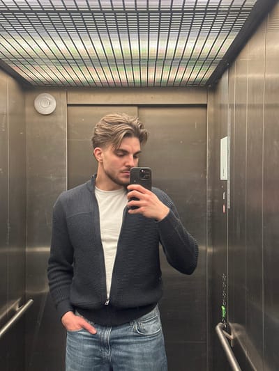 A male individual, approximately 20-35 years of age, with light brown hair and a short beard, is captured taking a mirror selfie inside an elevator in Volketswil, Switzerland. He is dressed in a white crew-neck t-shirt, an unzipped dark blue waffle-knit zip-up cardigan, and blue denim jeans. His left hand is positioned in his jeans pocket, while his right hand holds a dark-colored smartphone, oriented to photograph his reflection. The elevator interior features reflective brushed stainless steel panels on the walls. The ceiling comprises a grid structure with linear lighting elements behind it, casting soft green and yellow hues. A circular white fixture is visible on the upper left wall, and a vertical control panel with green indicator lights is present on the right wall. A metallic handrail is visible in the lower right section of the image. The subject's gaze is directed towards the smartphone screen, not directly at the camera.
