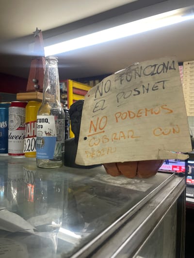 The image depicts a sign written in Spanish that reads "NO FUNCIONA EL POSNET. NO PODEMOS COBRAR CON DEBITO," meaning "THE POSNET IS NOT WORKING. WE CANNOT CHARGE WITH DEBIT." This indicates a business is unable to process debit card payments. The sign is placed among empty beer bottles, including Corona Cero, Budweiser, and other brands, stacked on a surface that appears to be part of a display or storage area. The environment seems to be a store or establishment in Vicente López, Argentina, based on the labeling on the Corona Cero bottle indicating production in Argentina.