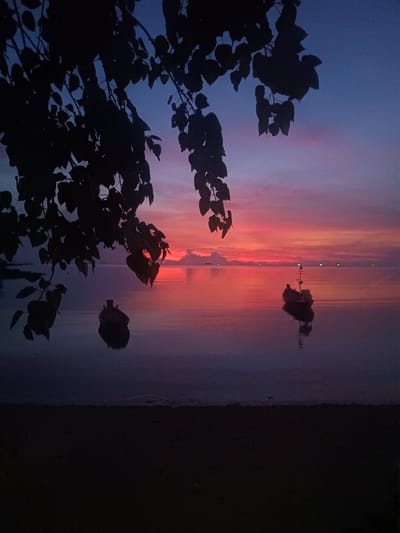The image presents a twilight or sunset scene at Baan Wok Tum, Thailand. The sky is a gradient of blue, purple, and vibrant red hues, reflected in the calm water of the sea or bay. Two small boats are visible on the water, appearing as dark silhouettes against the colorful reflection. Silhouetted tree branches and leaves hang in the foreground, adding depth to the scene. The foreground also has a small portion of beach or shoreline. Lights shine on the horizon line.
