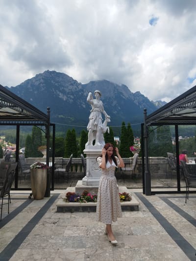 The image shows a young woman standing in front of a classical sculpture of a woman and a deer. Behind the sculpture, a large mountain range with a cross at its peak dominates the background. The scene appears to be an outdoor terrace with tables and chairs, suggesting it might be a restaurant or cafe. The woman is wearing a long, floral-print dress and white heels. She is touching her hair, looking towards the camera. The location is identified as the Bușteni City Hall in Bușteni, Romania, which is known for its panoramic views of the Bucegi Mountains. The sky is cloudy, with dramatic cloud formations adding to the scenic grandeur. The overall impression is one of serene beauty, with the woman as the focal point against the majestic natural backdrop.